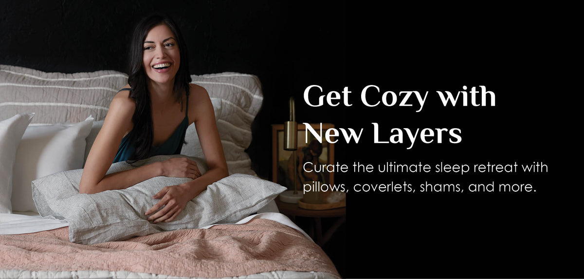 Image of a woman sitting up in bed smiling with Heritage Pillow Shams behind her, a Wave coverlet covering her, and a pillow with a pinstripe Hemp Pillowcase in her lap. A caption to the right of her reads: "Get Cozy with New Layers, Create the ultimate sleep retreat with pillows, coverlets, shams, and more."