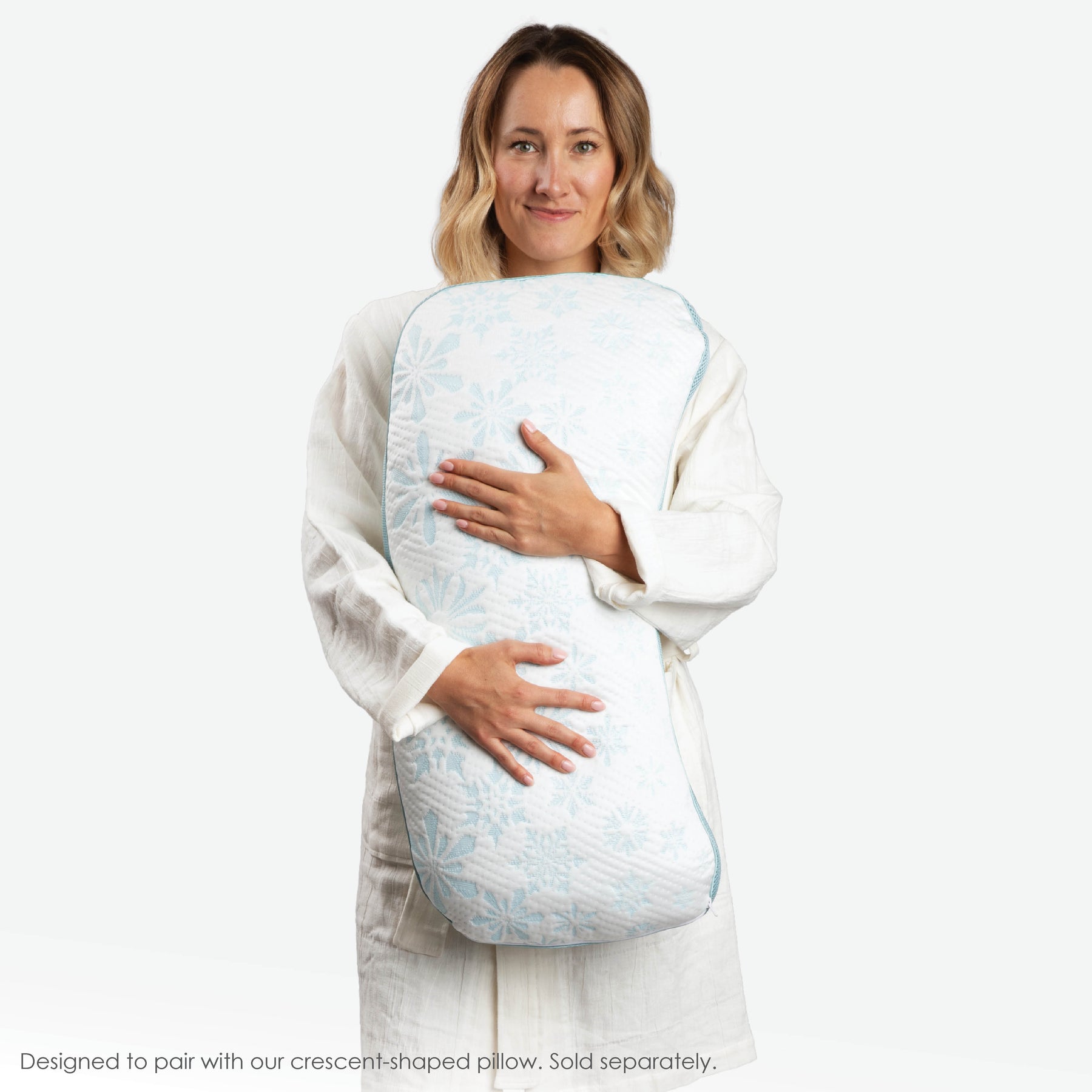 Image of a woman in a Dr. Weil Featherweight Robe standing up and hugging the Cooling Crescent Shredded Foam Pillow against her. At the bottom of the image, a caption says "Designed to pair with our crescent-shaped pillow. Sold separately."