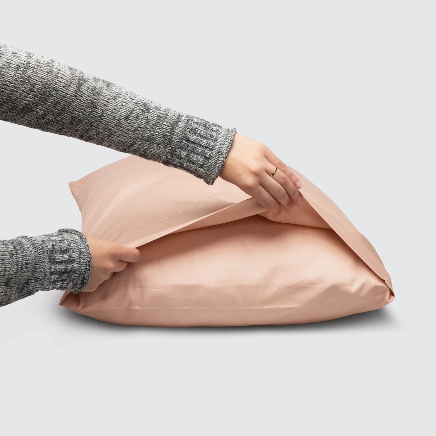 Image of a Blush Pink Weightless Cotton Pillowcase on a pillow with a hand lifting up the edge to showcase the enveloping design