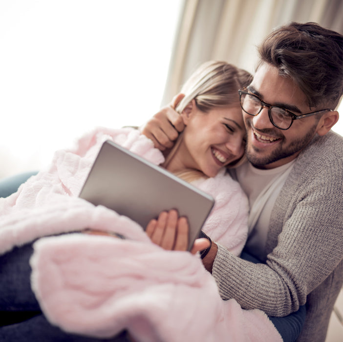 Image of a woman and man sitting together and laughing at a tablet as the woman is wrapped in a Soft Pink blanket 