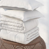 Image of a pile of bedding on top of a brown table. The bedding shown includes (from top to bottom): 2 White Pillow Shams, a neatly folded White Duvet Cover + Cooling, and a neatly folded Cooling Duvet Insert 